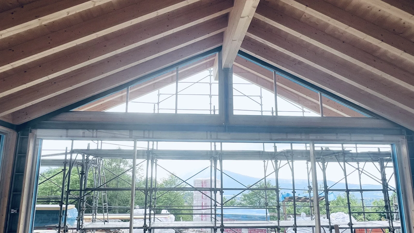 La grande vetrata sul panorama del Lago di Santa Croce e del Nevegal: il cantiere di Acciaio, legno e aria visto dall'interno, con il timpano in legno lamellare e i ponteggi che incorniciano la vista