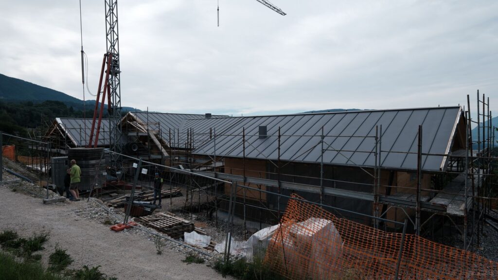 Vista d’insieme del cantiere con la casa e il tetto in lamiera scura completato, montagne sullo sfondo.