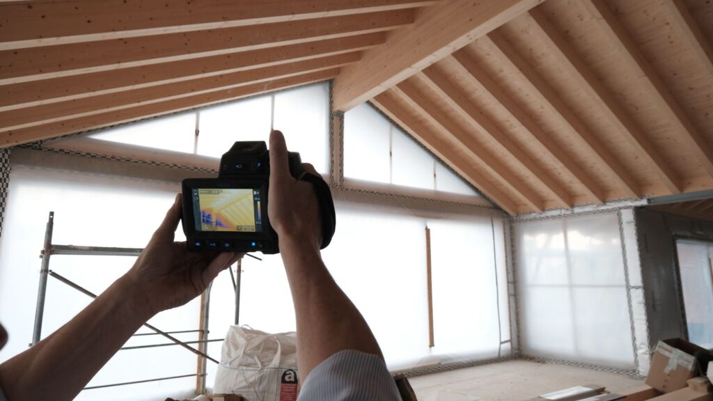 Mani che reggono una termocamera puntata verso il nodo tra tetto in legno e parete durante la prova di tenuta