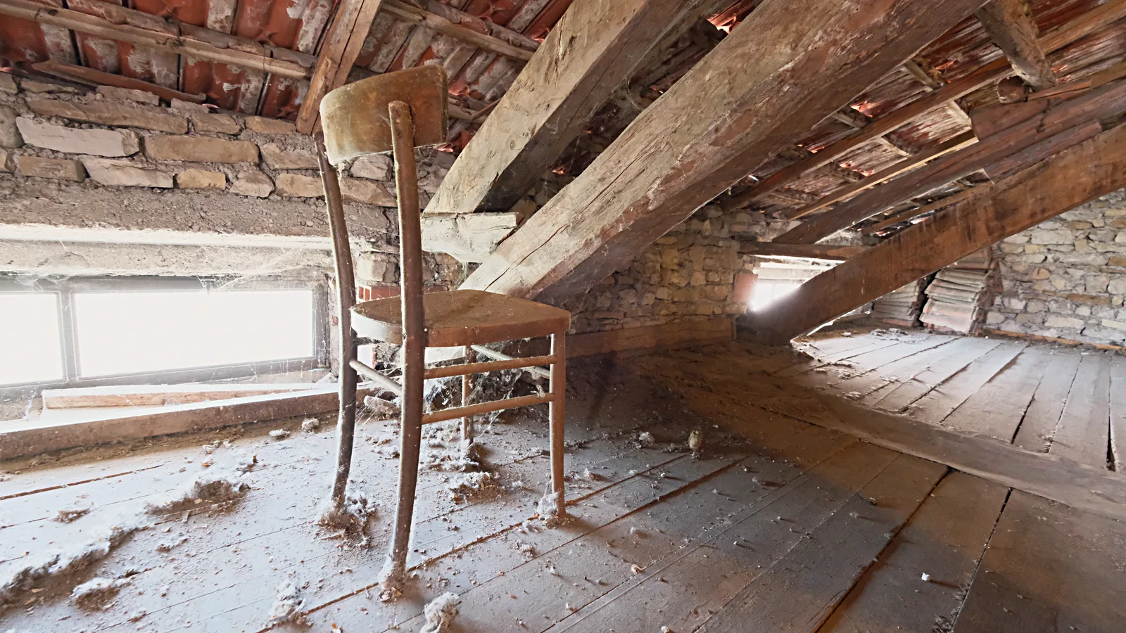 Una vecchia sedia impagliata rimasta sola nel granaio polveroso della casa ereditata, con la luce che entra dalla finestra bassa sotto il tetto in legno.