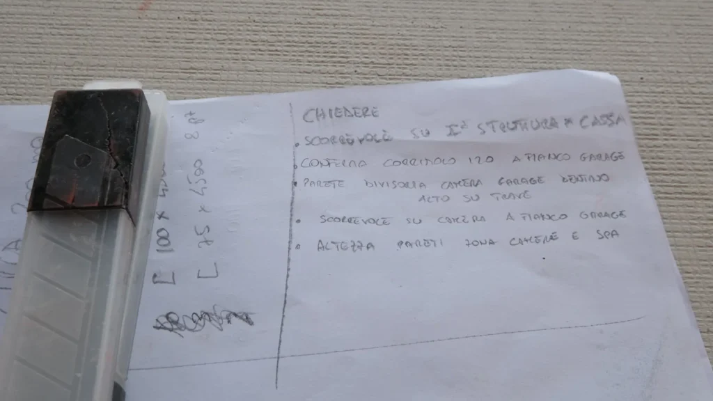 Foglio di appunti manoscritti di cantiere con un elenco di verifiche, sopra un cutter.