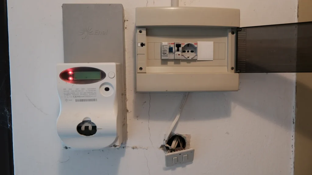 Un contatore elettrico e un quadretto con interruttori su una parete, accanto a una scatola di derivazione aperta nel muro con i fili a vista.
