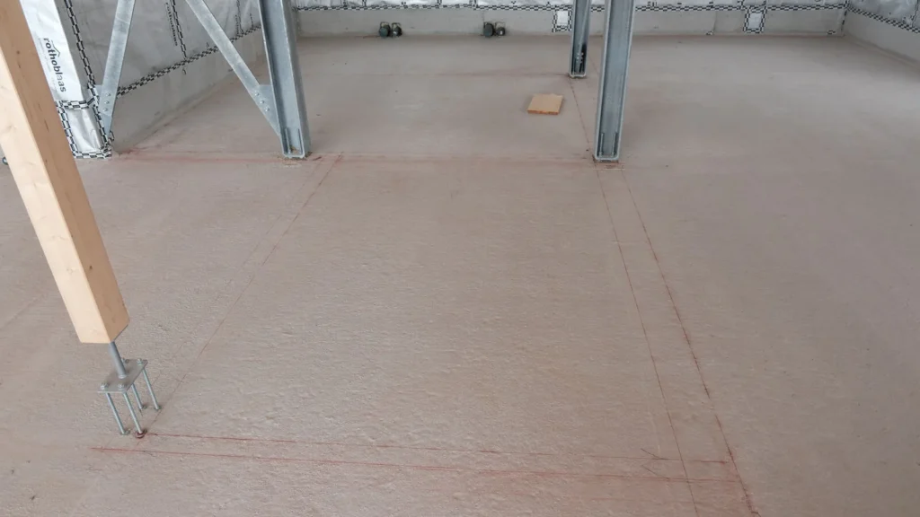 Linee rosse di tracciamento sul massetto di un cantiere, con un pilastro in acciaio e uno in legno.