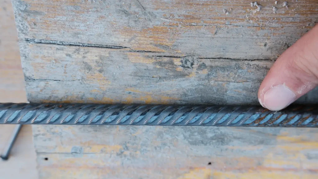 Un dito indica dall'alto le nervature a mezzaluna di una barra d'armatura appoggiata su una tavola di legno.