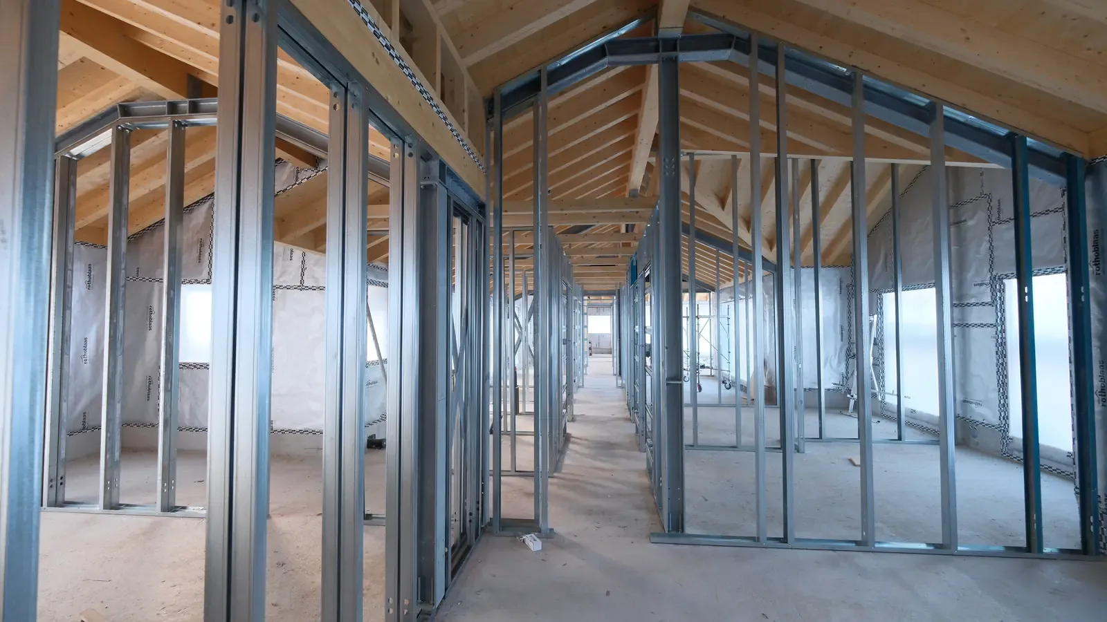 Interno di una casa in costruzione con struttura a montanti in acciaio e tetto in travi di legno a vista.