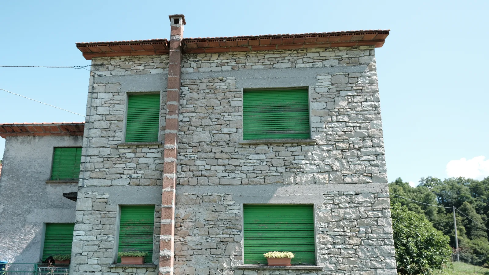 Casa in pietra a due piani nel Bellunese con gli scuri verdi chiusi e l'erba alta davanti alla facciata.