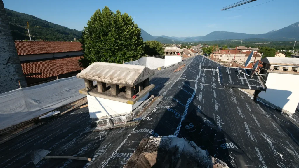 Falda di tetto coperta da membrana nera traspirante con due comignoli, tetti e montagne sullo sfondo.