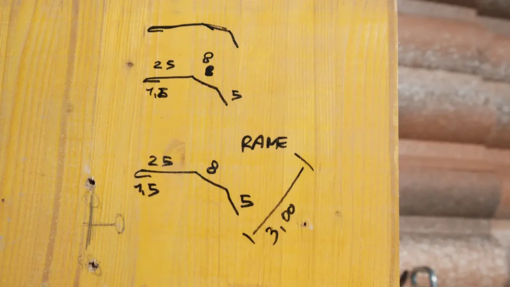 Schizzo a matita su tavola di legno gialla con le misure di un profilo di lattoneria in rame, accanto a tegole accatastate.