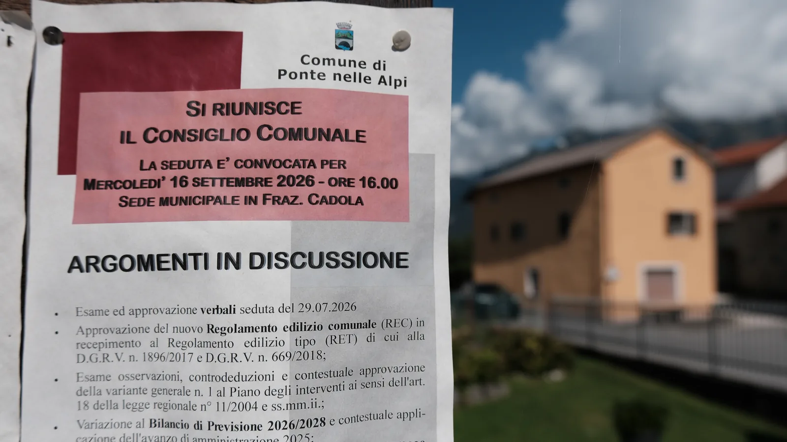L'avviso di convocazione del Consiglio comunale di Ponte nelle Alpi del 16 settembre 2026 appeso alla bacheca, con l'ordine del giorno; sullo sfondo, sfocata, una casa.