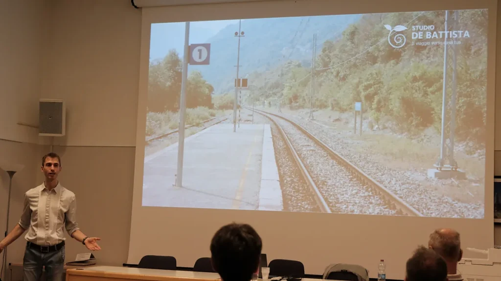 Lewis Dal Magro parla a braccia aperte davanti allo schermo con la foto di una stazione e i binari; in primo piano il pubblico di spalle.