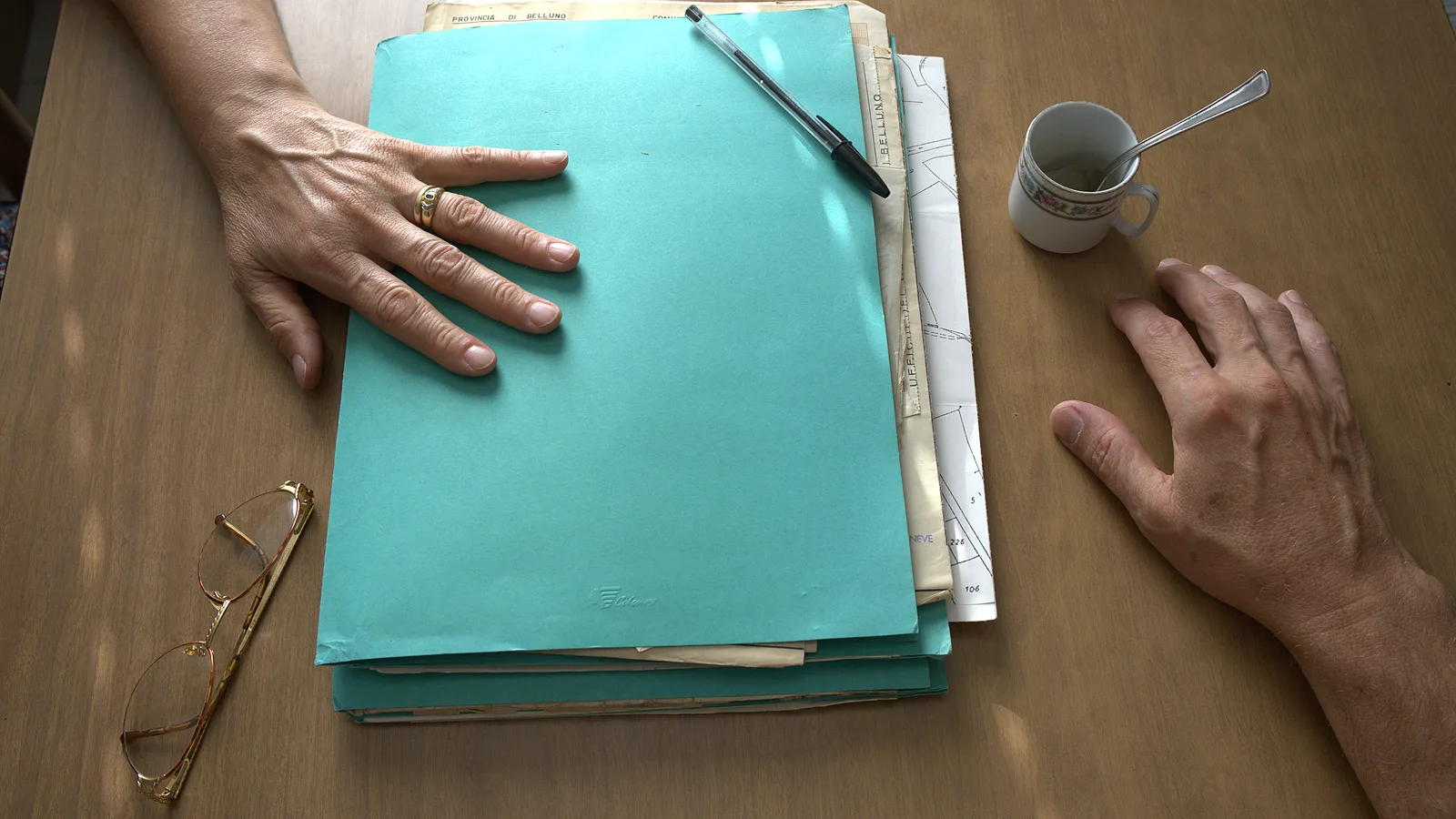Due mani ai lati di una cartellina di documenti chiusa su un tavolo di cucina, con una penna appoggiata sopra, occhiali da lettura e una tazzina di caffè.